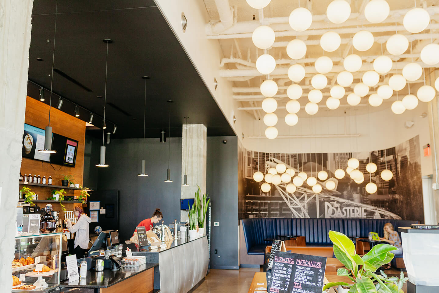 The inside of the Woodside Roasterie Café location. There is a cash register, beautiful chandelier lighting, and plant lift all around.