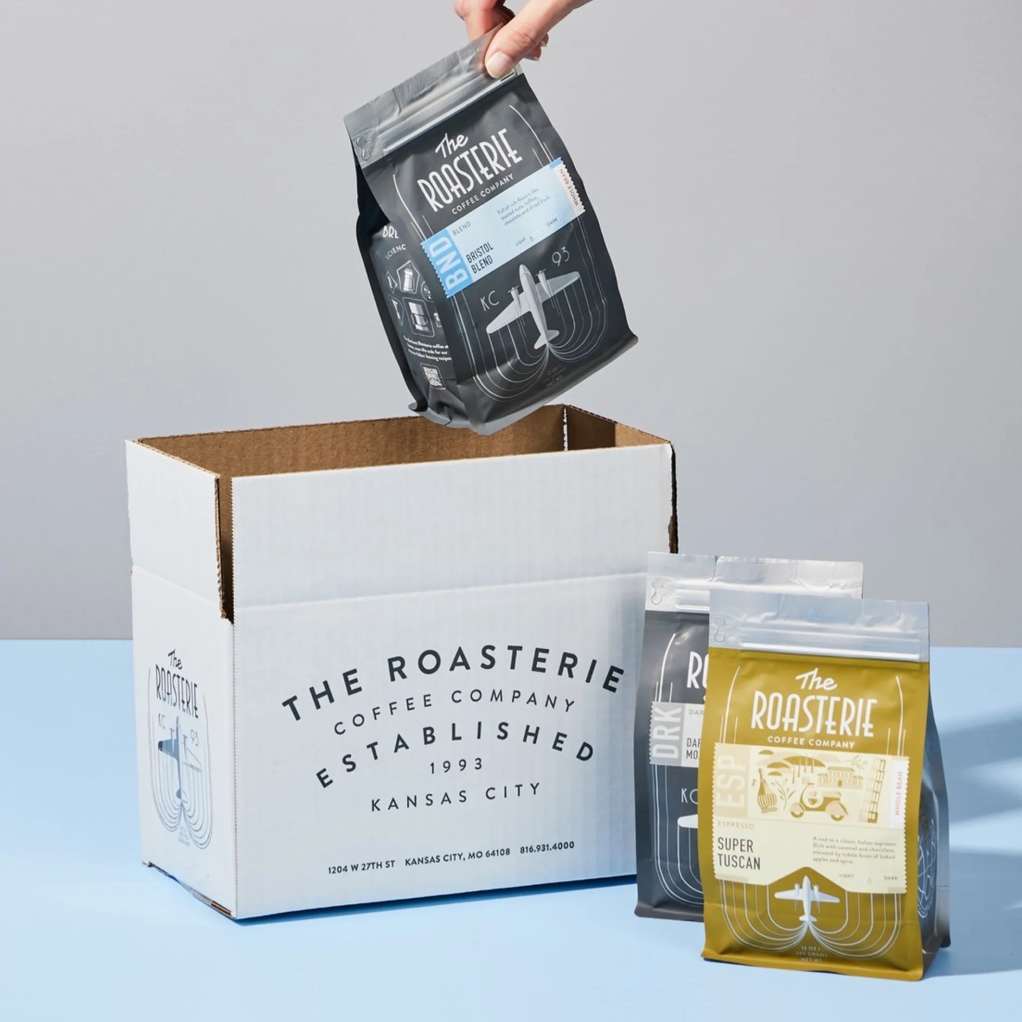 The Roasterie coffee packaging with a hand lifting a bag, displayed on a light blue background.