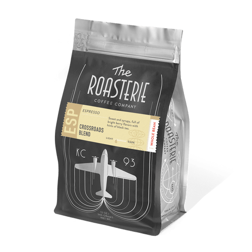 A black coffee bag of Crossroads Espresso by The Roasterie featuring an art deco-style airplane design in silver, with a gold label highlighting the blend name and tasting notes on a resealable package.